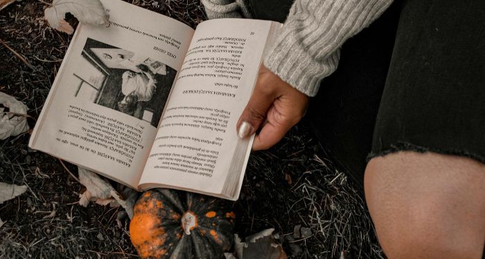 a brown skinned hand holding open a book on the ground there is a pumpkin under the book.jpg.optimal