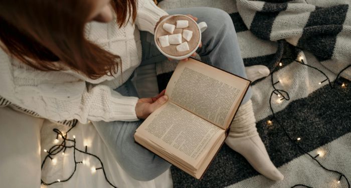 fair skinned red haired person sitting cross legged and reading with a cup of hot chocolate.jpg.optimal