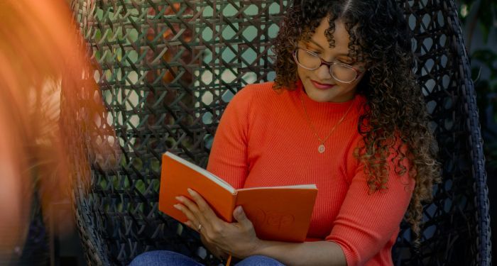 a light tan skinned woman with long curly hair and an orange shirt reading a book in a wicker chair