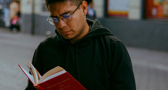 a tan skinned man with glasses reading a book on a sidewalk.jpg.optimal